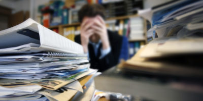 Stressad man på kontoret med hög av dokument och pärmar på bordet.